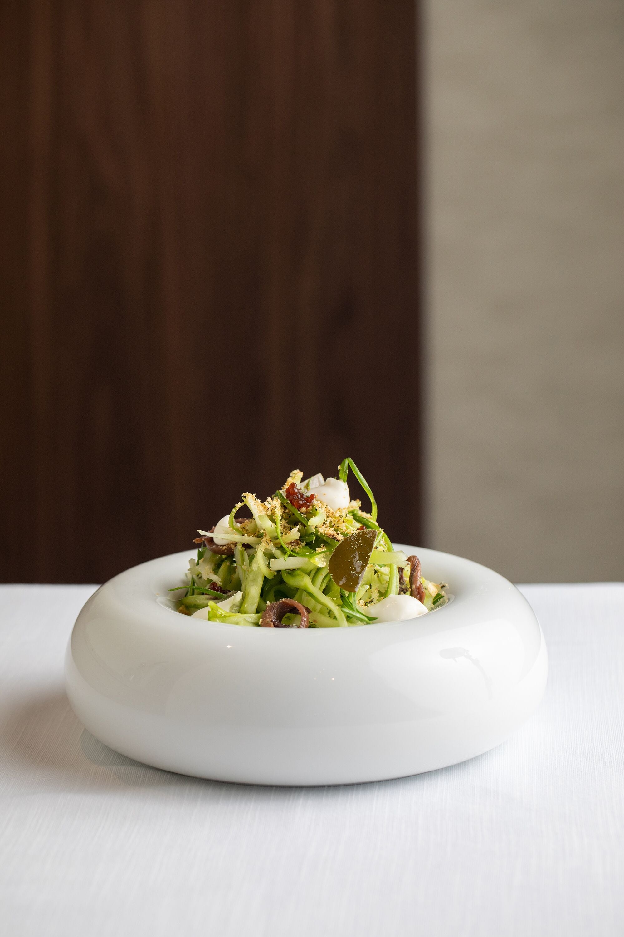 A white plate with a salad on a white table against a wooden background.