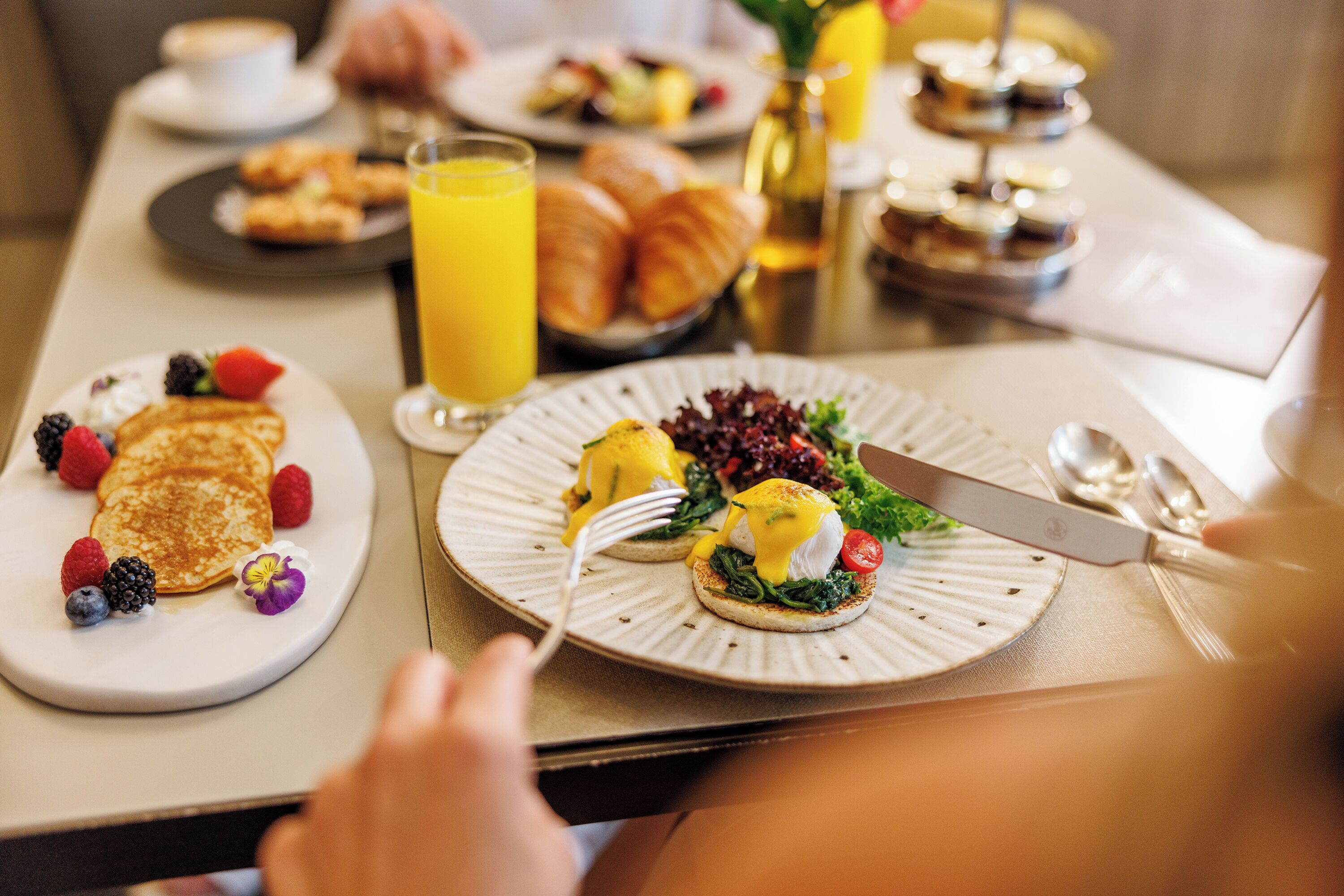 Un desayuno con platos de panqueques, huevos Benedictinos, cruasanes y un vaso de jugo de naranja.