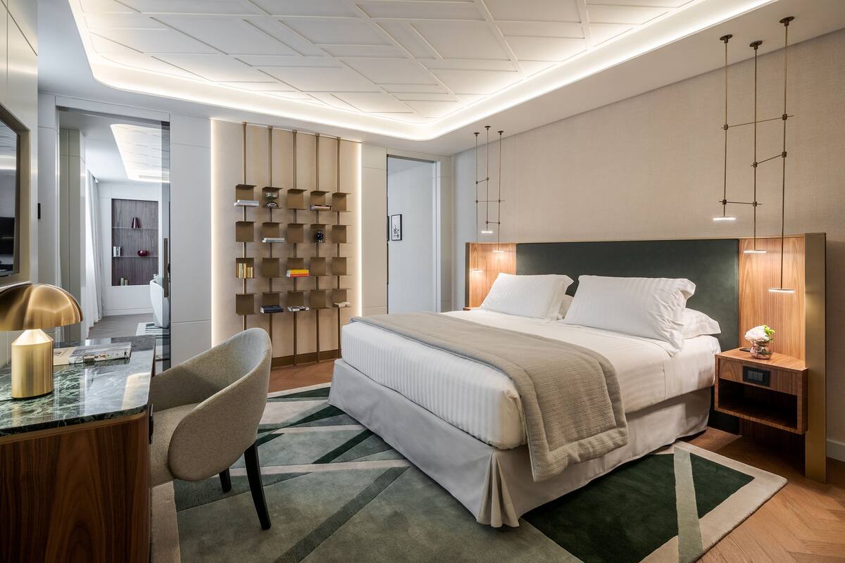 Ein modernes Hotelzimmer mit einem großen Bett, einem Schreibtisch mit Lampe, einem Sessel und einem Bücherregal.