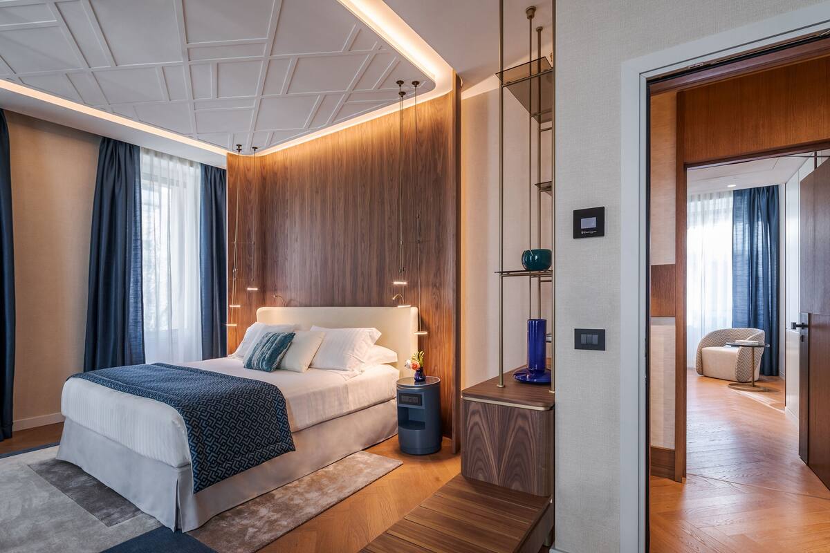 Ein modernes Hotelzimmer mit einem großen Bett, Holzwandverkleidung, einem Nachttisch mit Lampe und Blick in das angrenzende Zimmer.