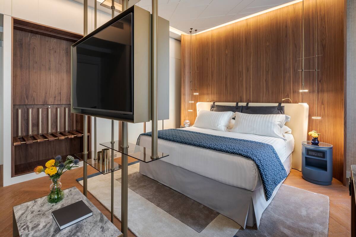 Ein modernes Hotelzimmer mit einem großen Bett, einem Flachbildfernseher an einer Glasstativ und einer Wand mit Holzpaneelen sowie einem Nachttisch mit Lampe.