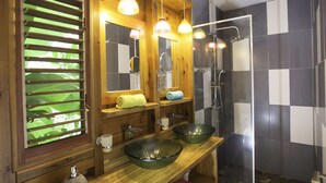 Casa de banho com toucador de madeira, duas tigelas de vidro verde, um espelho e área de chuveiro com soalho de pedra.