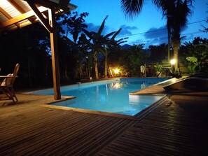 Una piscina con cubierta de madera y sillas de descanso, rodeada de follaje tropical e iluminación nocturna.