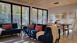 A modern living room with a dark blue sofa, a wooden dining table with chairs, and large windows offering a view of a forest.