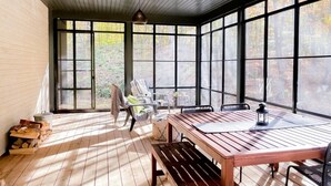 A sunlit room with a wooden table, chairs, and a large window overlooking a forest.
