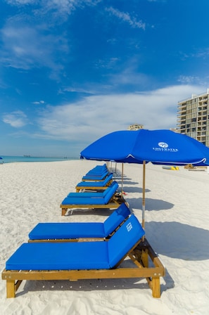 Une rangée de chaises longues bleues sous un parasol bleu, sur une plage de sable, avec un ciel dégagé et une jetée visible au loin.