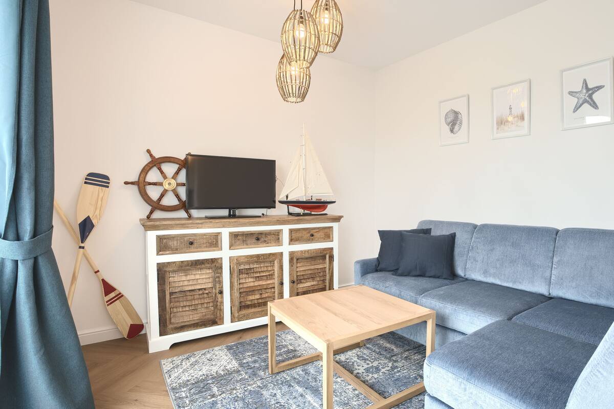 Ein modernes Wohnzimmer mit grauem Sofa, Holztisch und Fernseher auf einer Holzkommode. An der Wand hängen zwei Holzschläger und ein Schiffsrad.