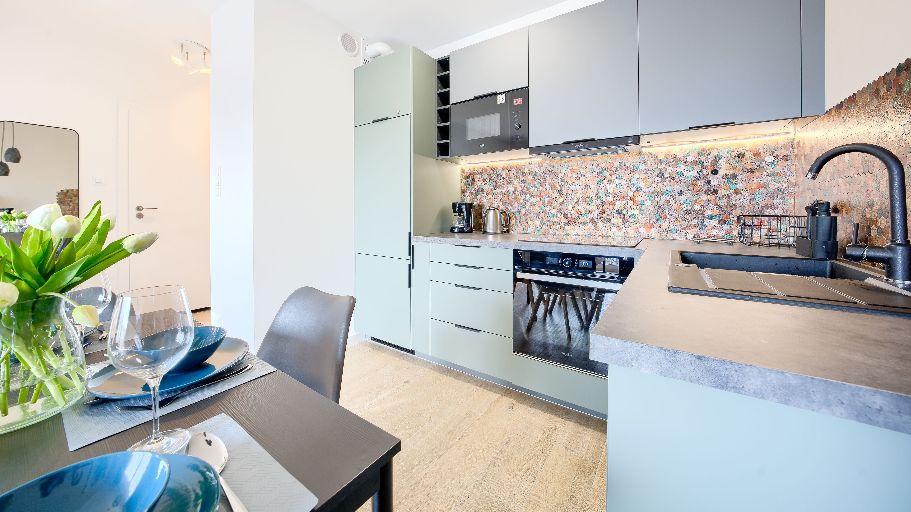 A modern kitchen with a microwave, stove, and sink. There is a dining area with a table set for two and a vase with flowers.