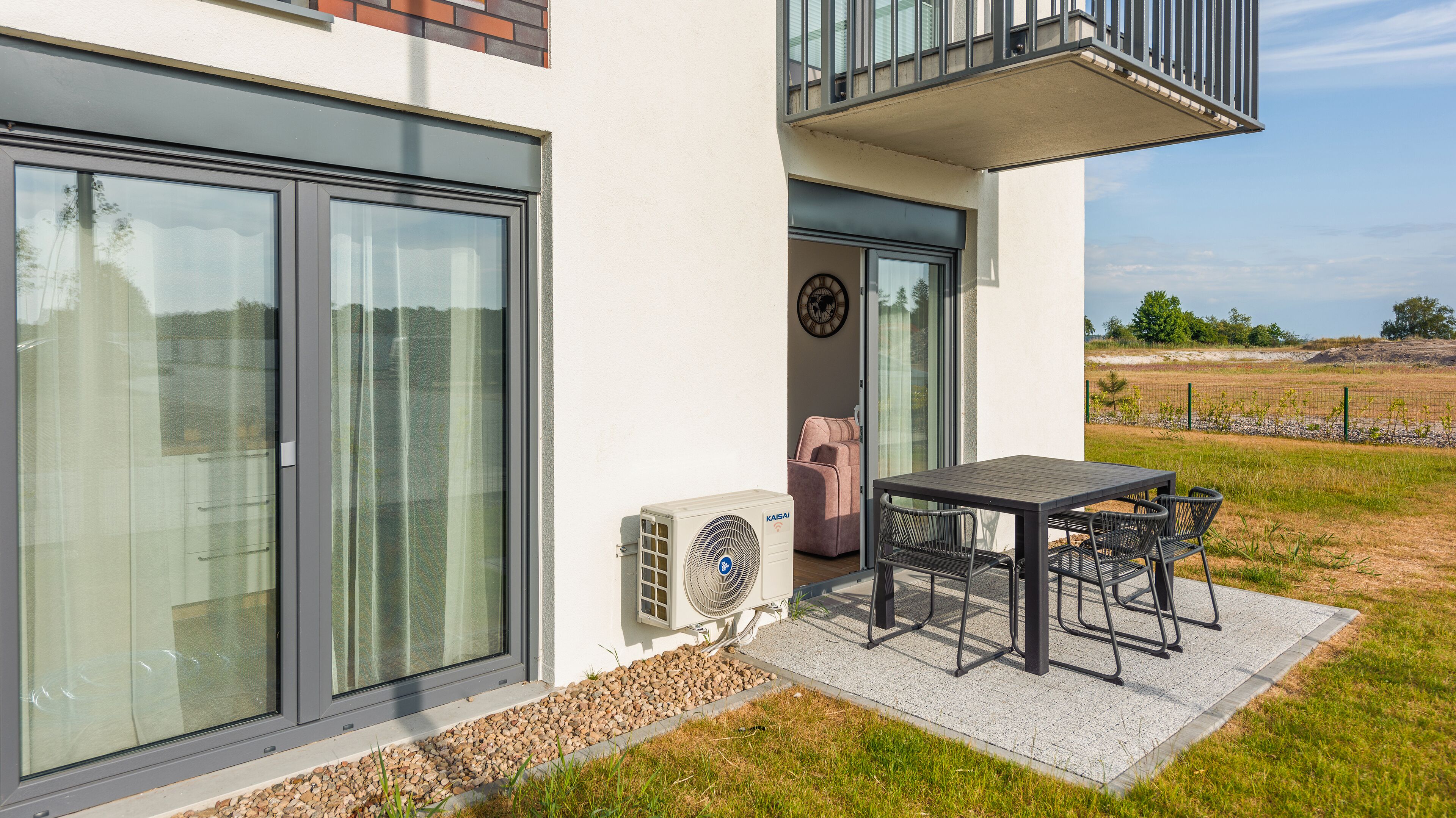 A modern house with a large sliding glass door, a small patio with a table and chairs, and a visible air conditioning unit.