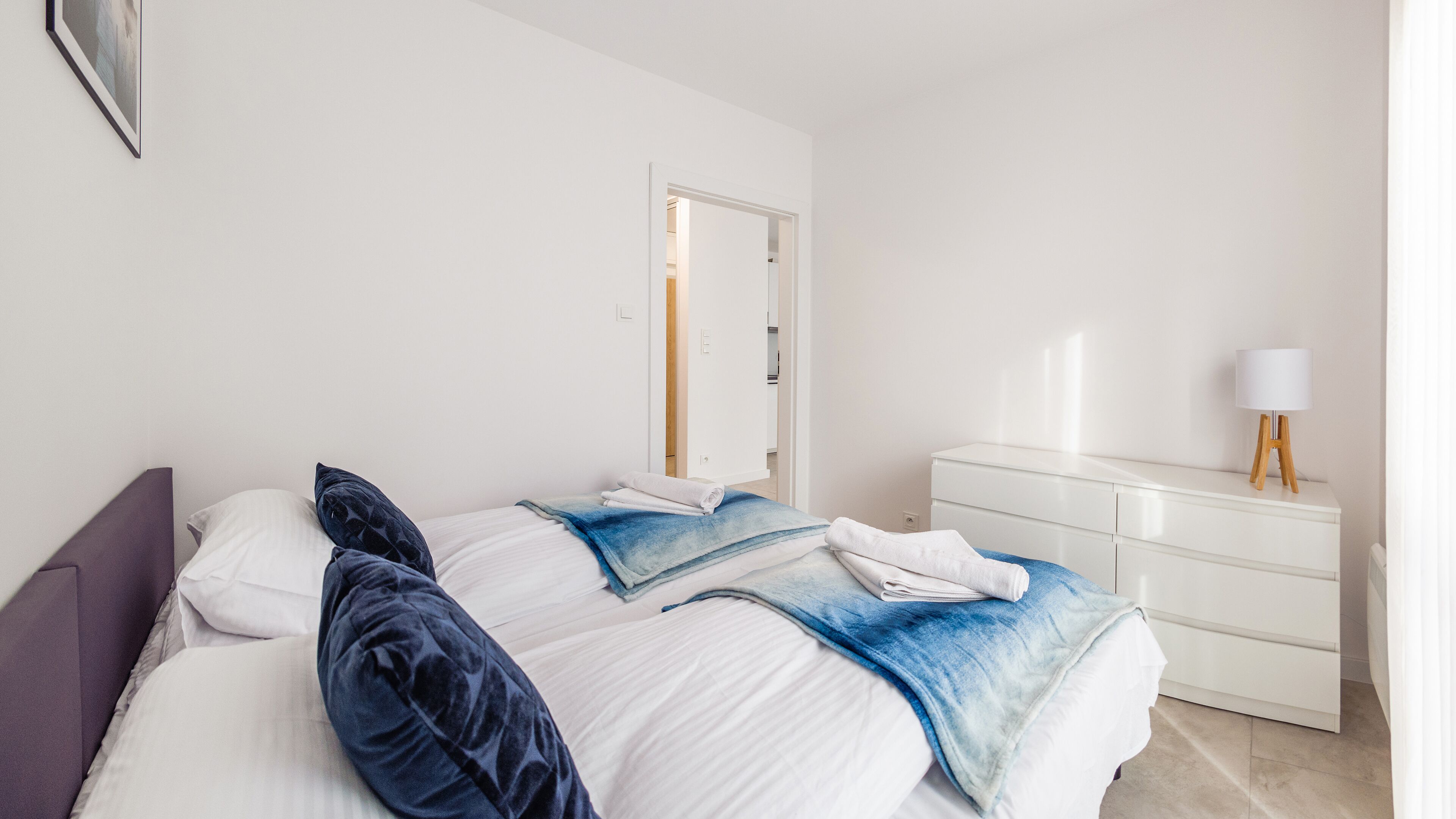 Un lit bien fait, avec des oreillers et une couverture, une commode blanche avec une lampe, et une porte qui donne sur une autre pièce.
