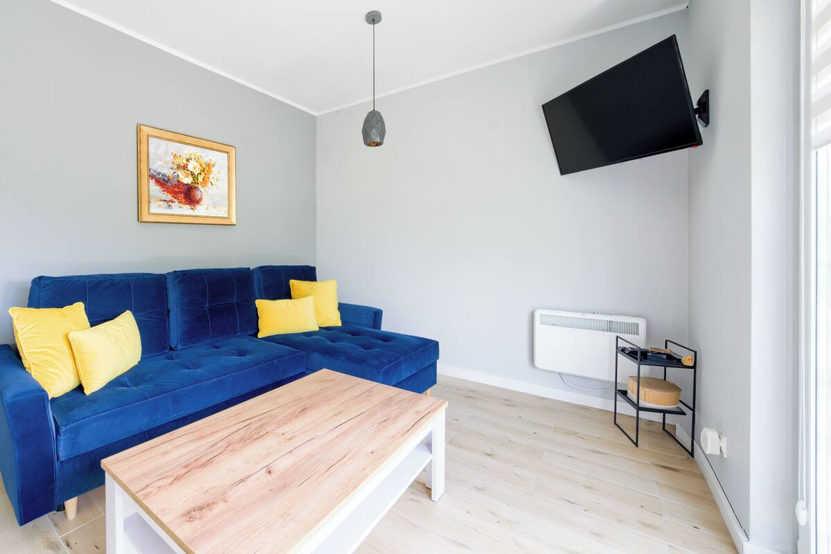 Ein Wohnzimmer mit blauem Sofa, gelben Kissen, einem Holztisch, einem gerahmten Bild, einem an der Wand befestigten Fernseher und einer Heizung.