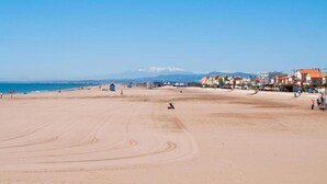 Una amplia playa arenosa con cielo azul despejado, una cadena montañosa distante y edificios con tejados rojos al fondo.