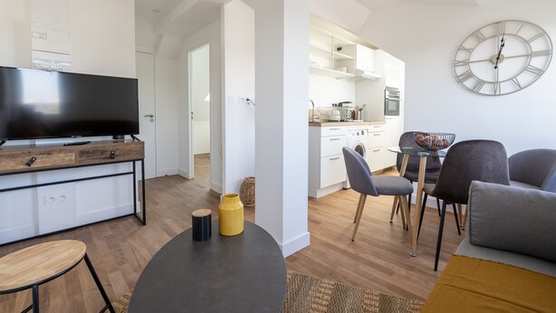 Un salon moderne avec un téléviseur à écran plat, une table à manger et un canapé. On y trouve également un coin cuisine avec des meubles blancs et une grande horloge au mur.