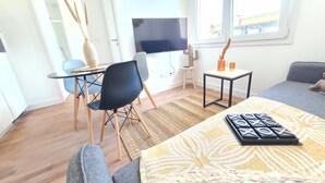 A modern living room with a sofa, a coffee table with a board game, a TV, and a window with a view of the outside. - Le Poulorio 7 -  T2 - Proche Gare * By Locly * (Lorient)