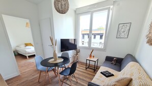 A modern living room with a sofa, a dining table with chairs, a TV, and a window with a view of buildings. - Le Poulorio 7 -  T2 - Proche Gare * By Locly * (Lorient)