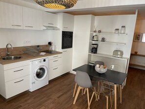 A modern kitchen with white cabinets, a built-in dishwasher, a washing machine, and a dining area with a black table and chairs. - LE POULORIO LOFT * Studio * RDC * ByLocly * (Lorient)
