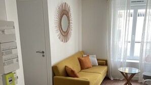 Un salon avec un canapé de couleur moutarde, un miroir rond, une petite table ronde et une fenêtre ornée de rideaux légers.