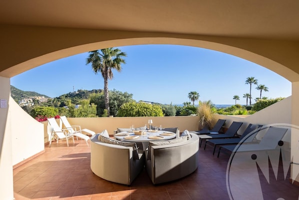 A sunlit outdoor seating area with a round table and lounge chairs, overlooking a coastal landscape with palm trees and hills.