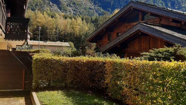 Une cabane en bois avec un toit en pente, entourée d’une haie bien entretenue et avec une chaîne de montagnes en arrière-plan.