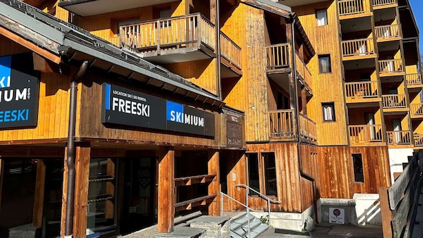 Ein mehrstöckiges Holzhaus mit Balkonen, Schildern für 'FREESKI' und 'SKI MIUM' sowie einer Skiverleih-Station.