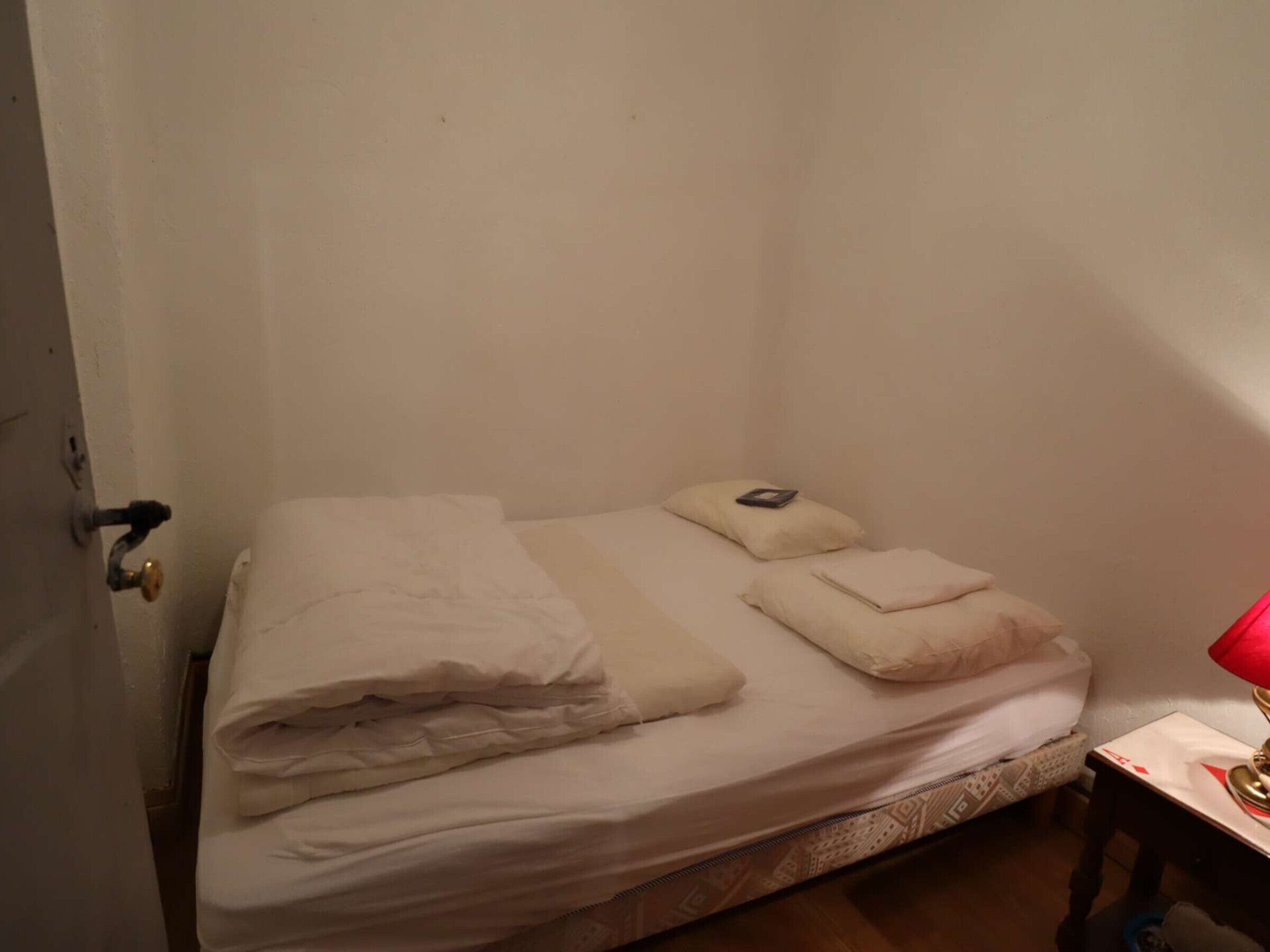 A small, single-bed room with a door on the left, a bedside table with a lamp, and a wall-mounted shelf.