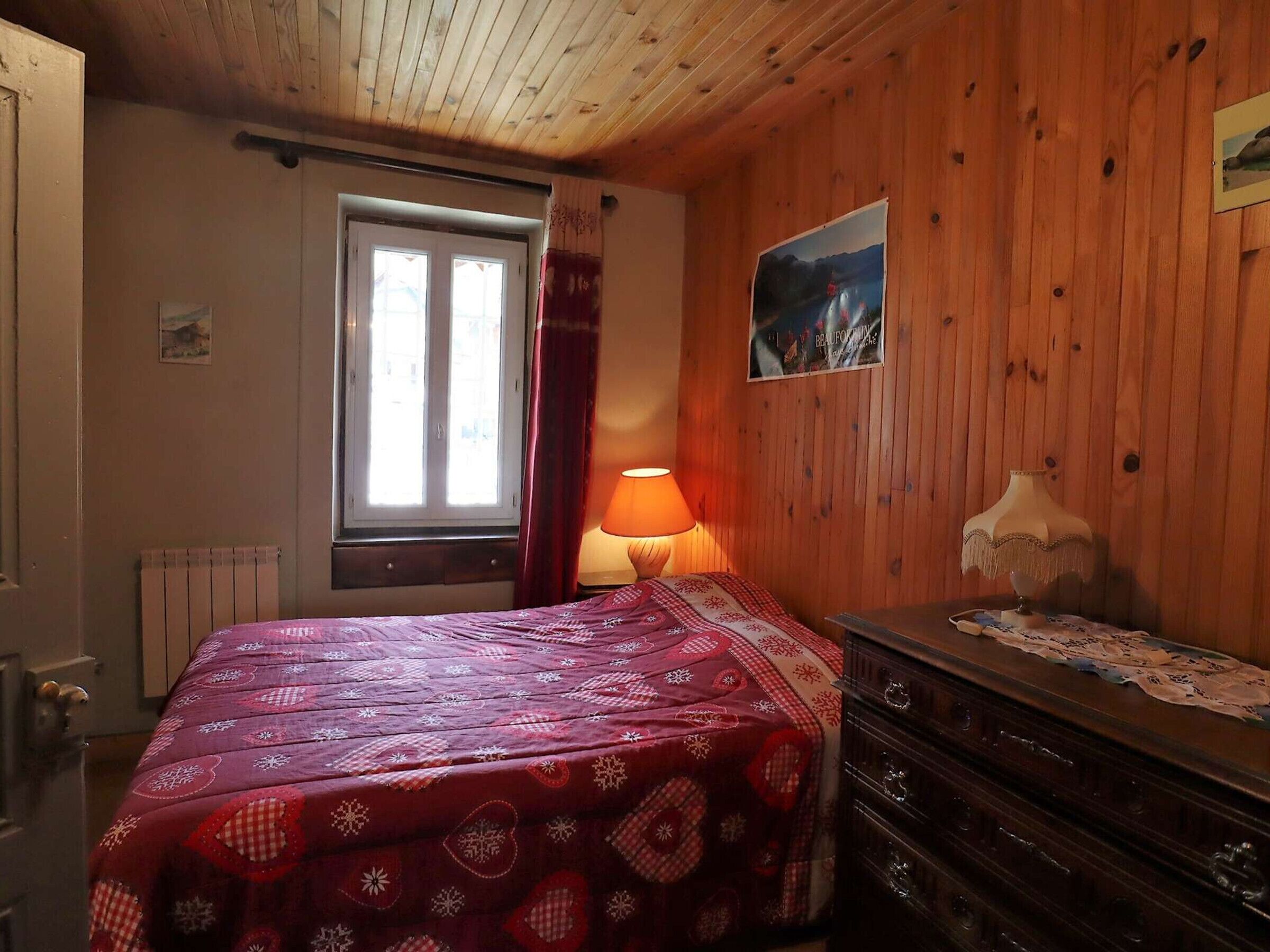 A cozy room with a wooden ceiling, a bed with a red and white patterned bedspread, a dresser with a lace cover, a window with curtains, and a wall-mounted picture.