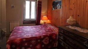 Una habitación acogedora con techo de madera, una cama con colcha de lunares rojos y blancos, un aparador con cubre de encaje, una ventana con cortinas y un cuadro colgado en la pared.