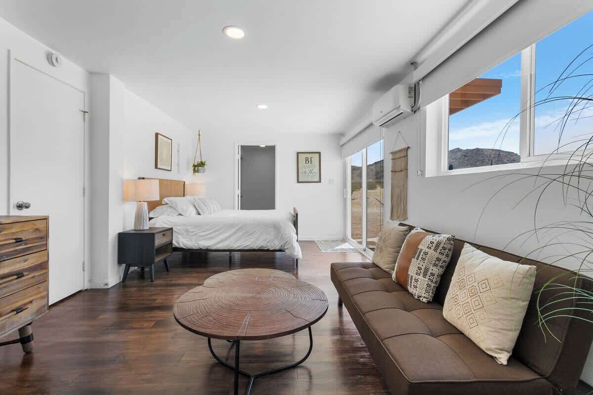 A modern bedroom with a large bed, a brown sofa, a round coffee table, and a view of the outdoors through large windows.