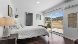 A bedroom with a bed, bedside lamp, and a view of a desert landscape through large windows. - 4BR w/ Hot Tub, Space X & Tesla Charger! (Joshua Tree)