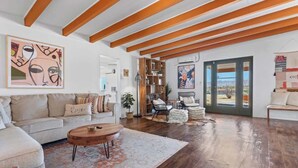 A spacious living room with a high ceiling, wooden beams, a large window, and a view of the outdoors. - 4BR w/ Hot Tub, Space X & Tesla Charger! (Joshua Tree)