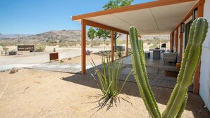 A desert landscape with a building featuring a covered patio, cacti, and a sign in the background. - 4BR w/ Hot Tub, Space X & Tesla Charger! (Joshua Tree)