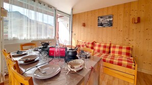 Ein Essbereich mit Tisch für zwei Personen, einer Holzsofa und Blick ins Freie durch ein Fenster mit durchsichtigen Vorhängen.