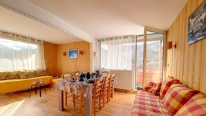 Ein gemütliches Zimmer mit Esstisch, Stühlen, einer Couch und einem großen Fenster mit Blick ins Freie.