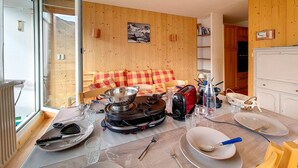 Ein gemütlicher Raum mit Holzwand, einer Couch mit karierten Kissen, einem Esstisch mit Tellern, Gläsern und Besteck sowie einem tragbaren Kochfeld.