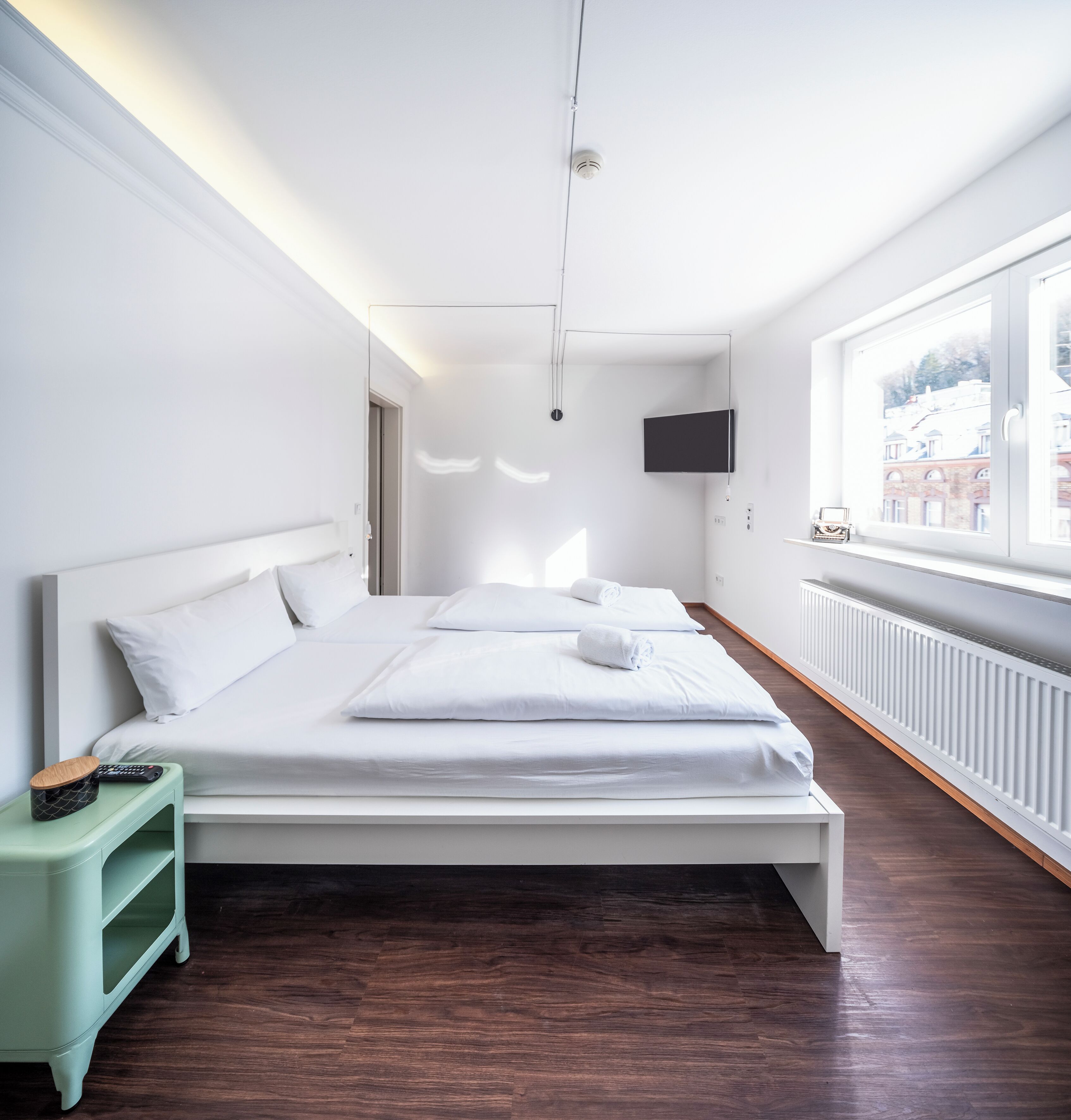 Una habitación de hotel moderna con una cama grande, una mesita auxiliar pequeña, un televisor montado en la pared y una ventana con vista a edificios.