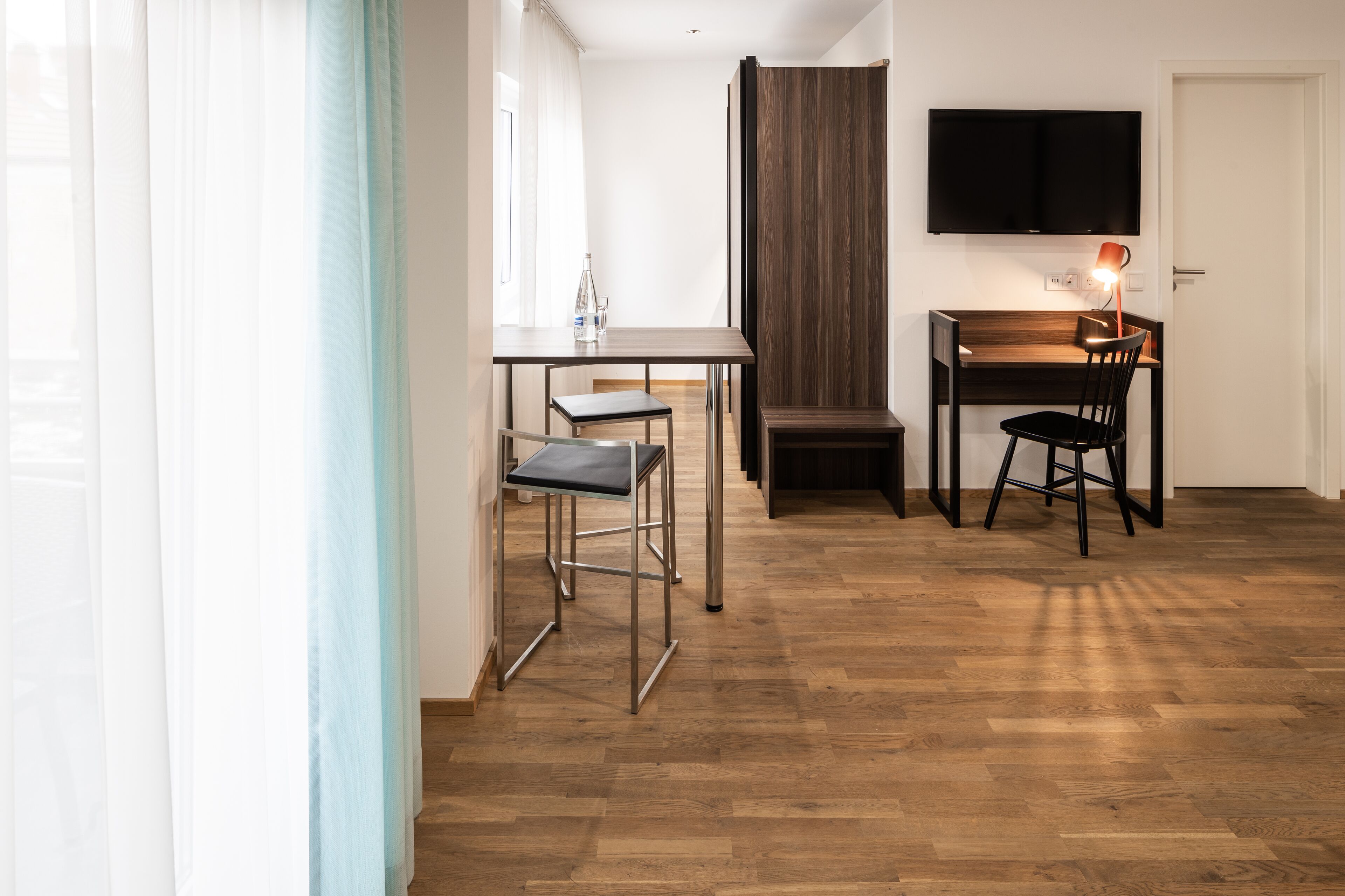 Una habitación moderna con piso de madera, una mesa de comedor con dos sillas, un televisor montado en la pared y un ventanal con cortinas.