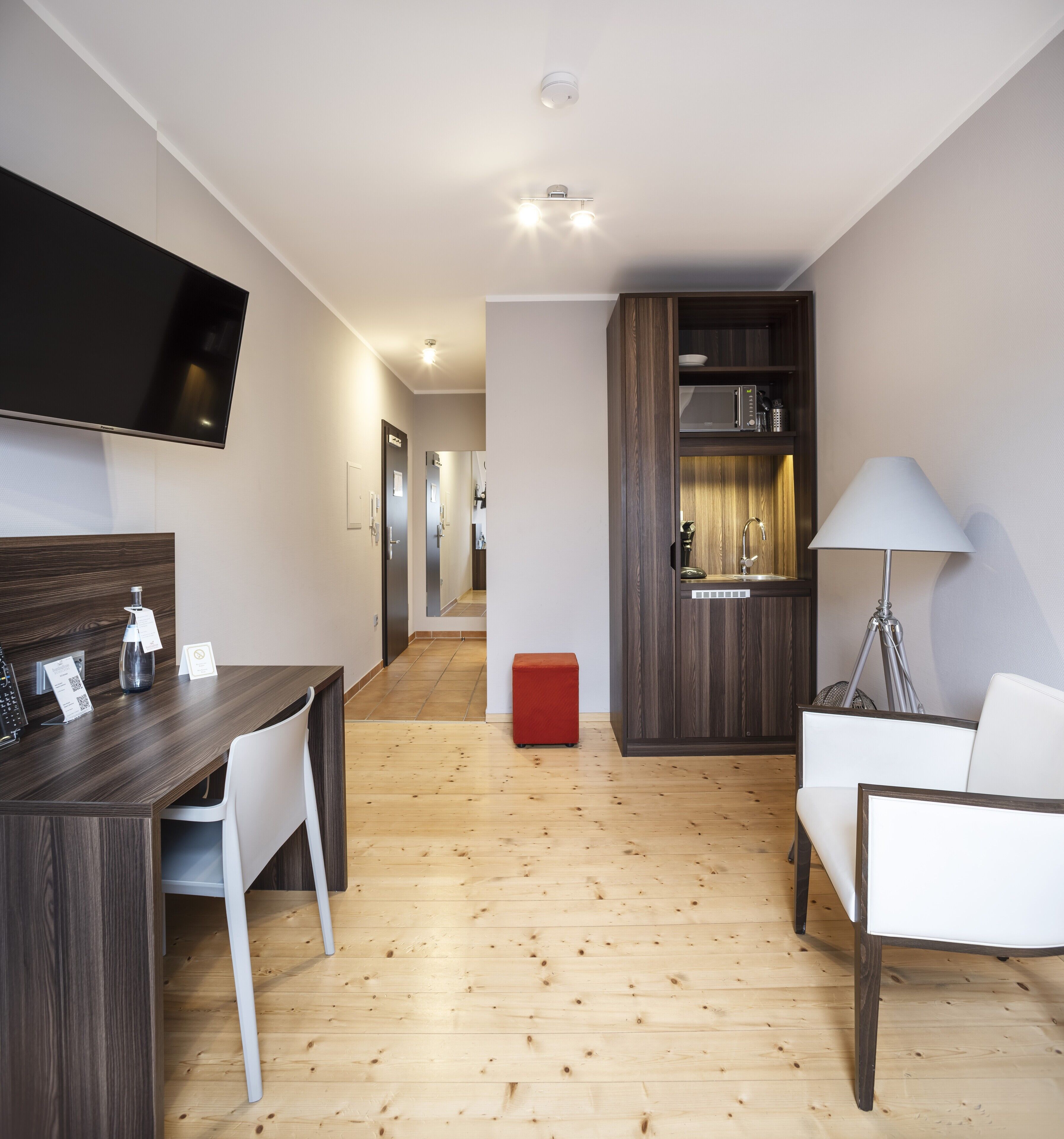 Une chambre d’hôtel moderne avec un plancher en bois, un coin repas comprenant une table et des chaises, un téléviseur fixé au mur, une kitchenette équipée d’un four à micro-ondes et un pouf rouge.