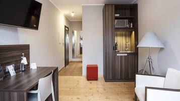 A modern hotel room with a wooden floor, a dining area with a table and chairs, a TV mounted on the wall, a kitchenette with a microwave, and a red ottoman.