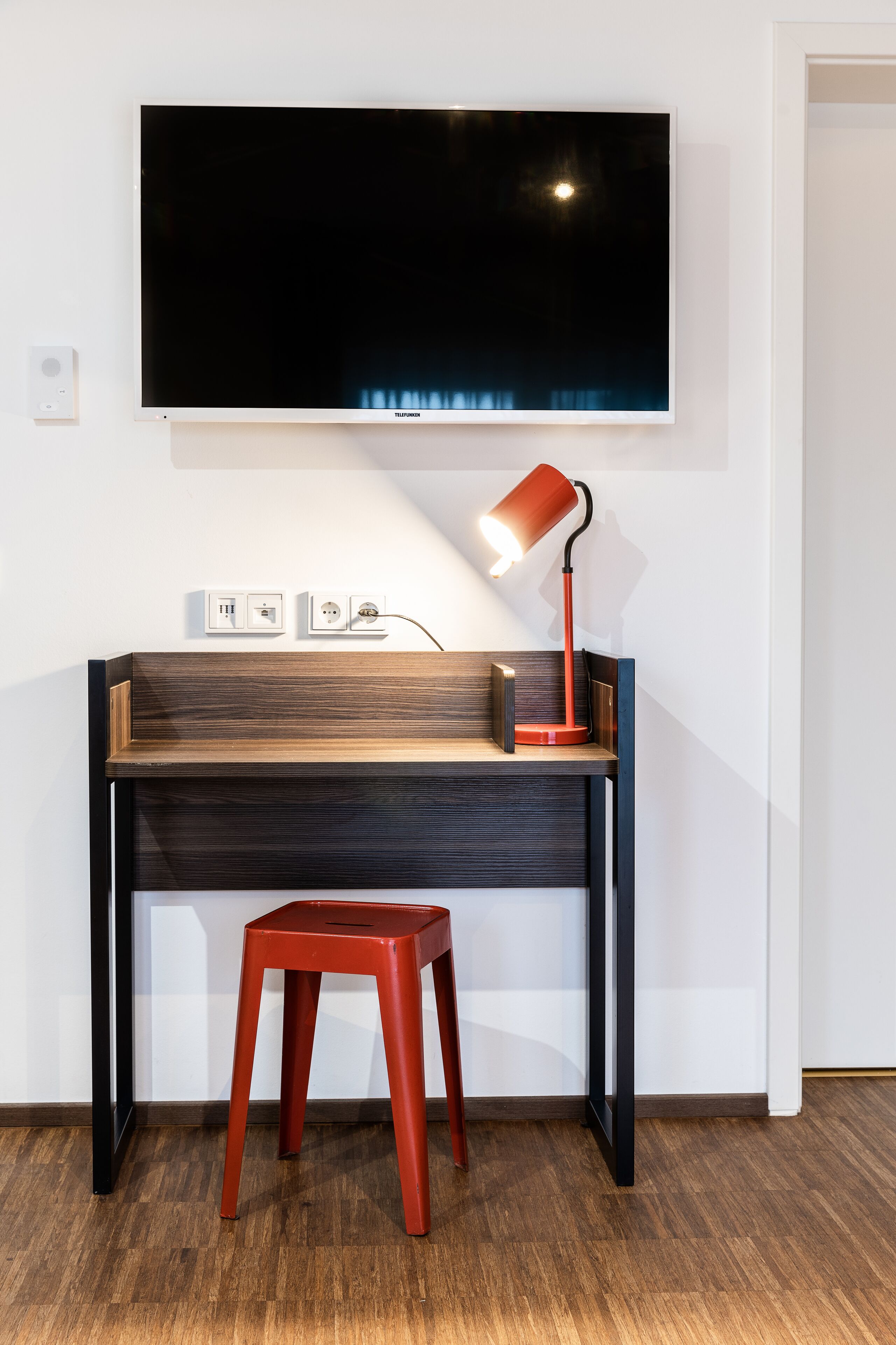 Ein moderner Arbeitsplatz mit einem Holztisch, einem roten Hocker, einem montierten Fernseher und einer roten Schreibtischlampe.