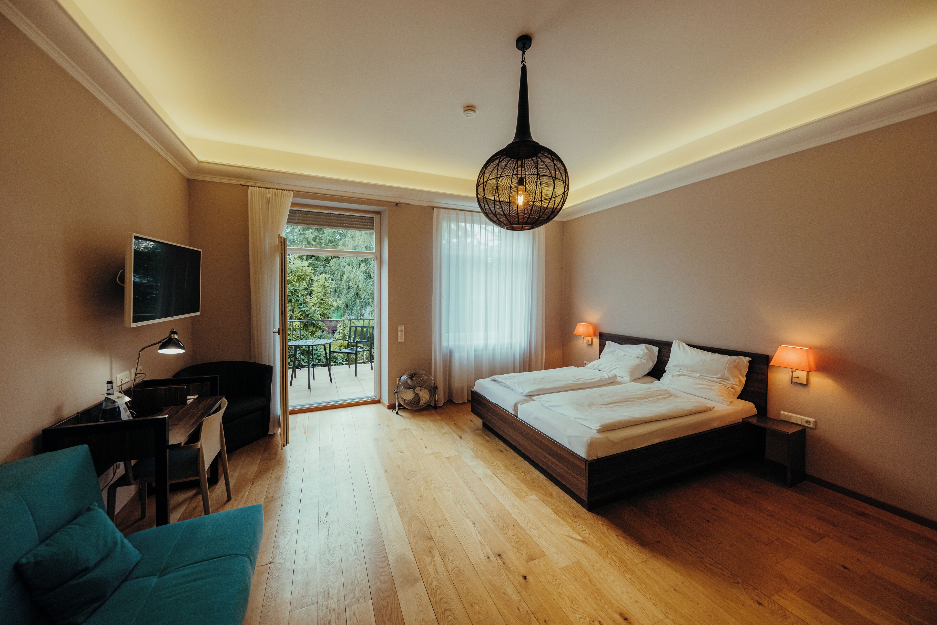 A bedroom with a bed, bedside lamps, a desk with a chair, a TV, and a view of greenery outside.