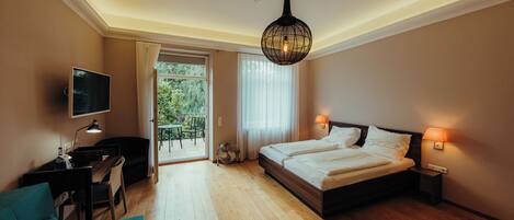 A bedroom with a bed, bedside lamps, a desk with a chair, a TV, and a view of greenery outside.
