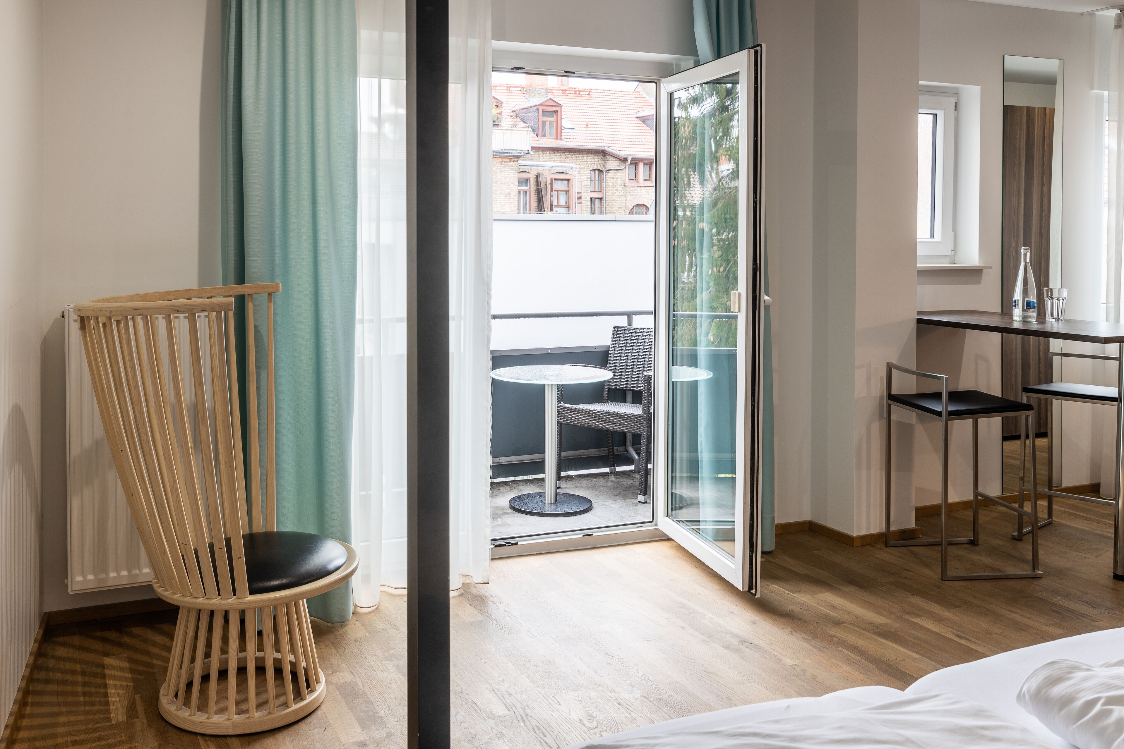 Ein Hotelzimmer mit einem Holzsessel, einem kleinen Tisch, einer Glastür, die auf einen Balkon führt, und einem Bett.