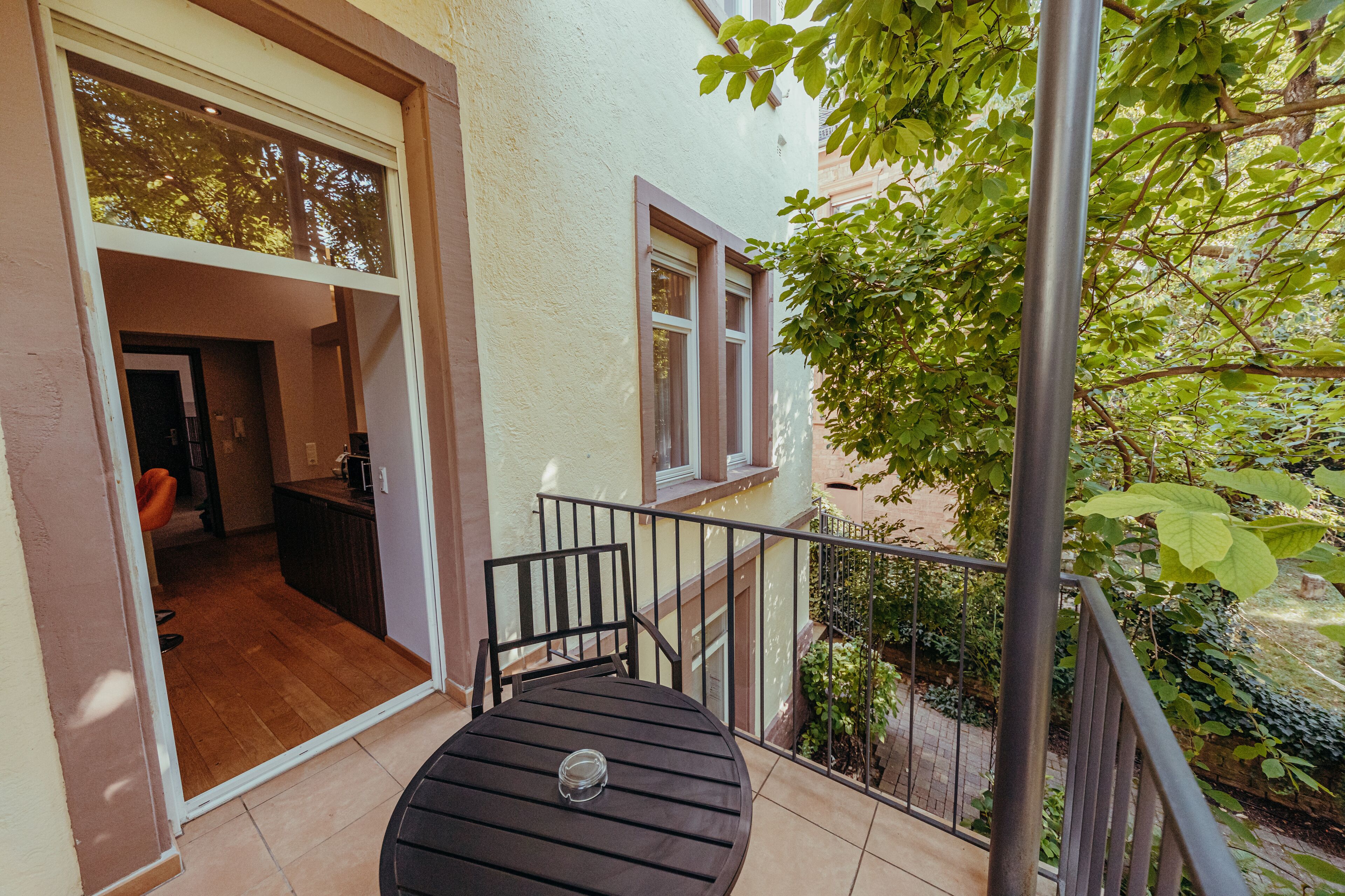 Een balkon met een tafel en stoelen, uitzicht op een tuin en een toegang tot een gebouw.
