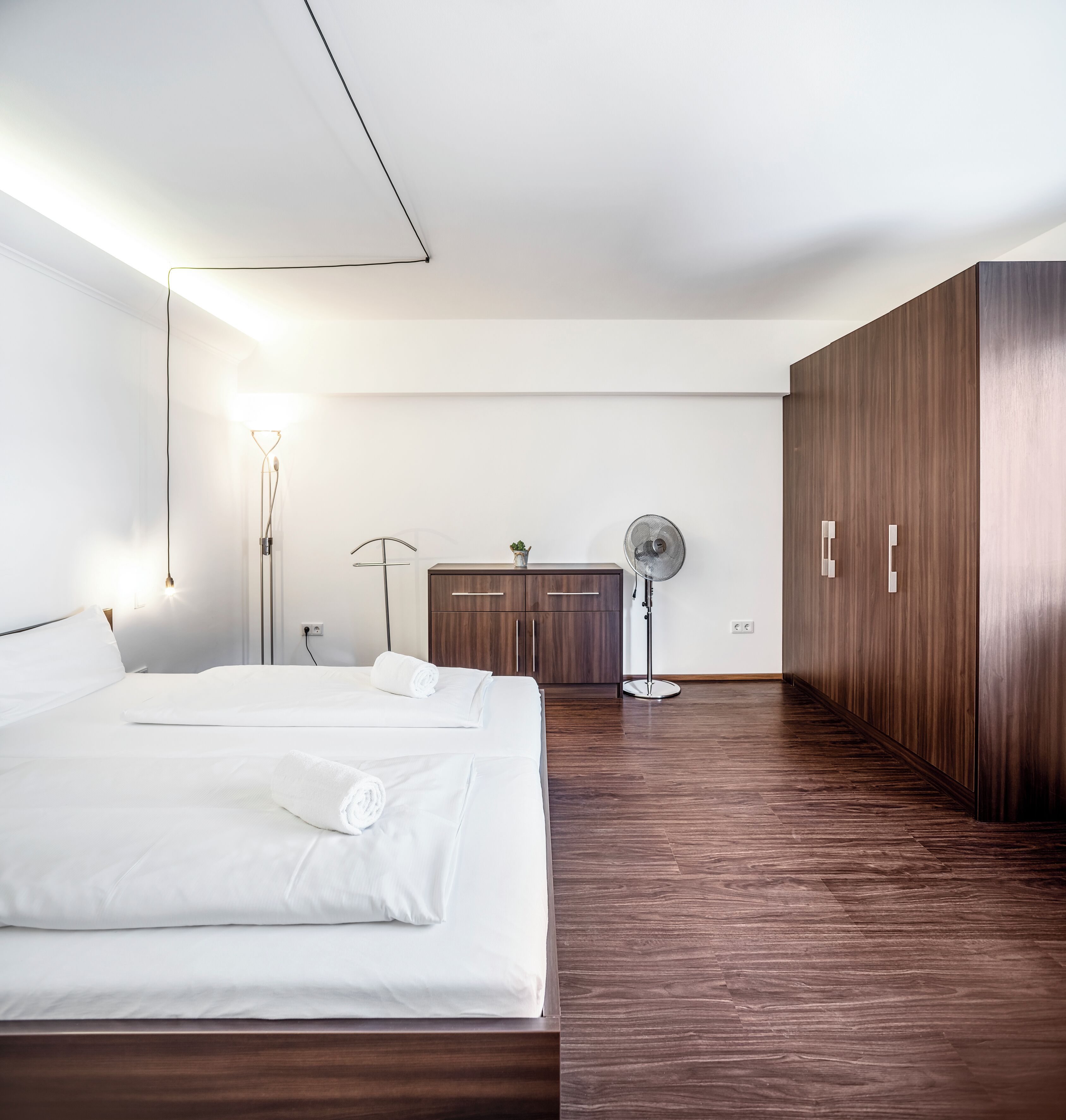 Ein modernes Hotelzimmer mit einem großen Bett, einem Holzschrank, einem Schreibtisch und einem Standventilator.