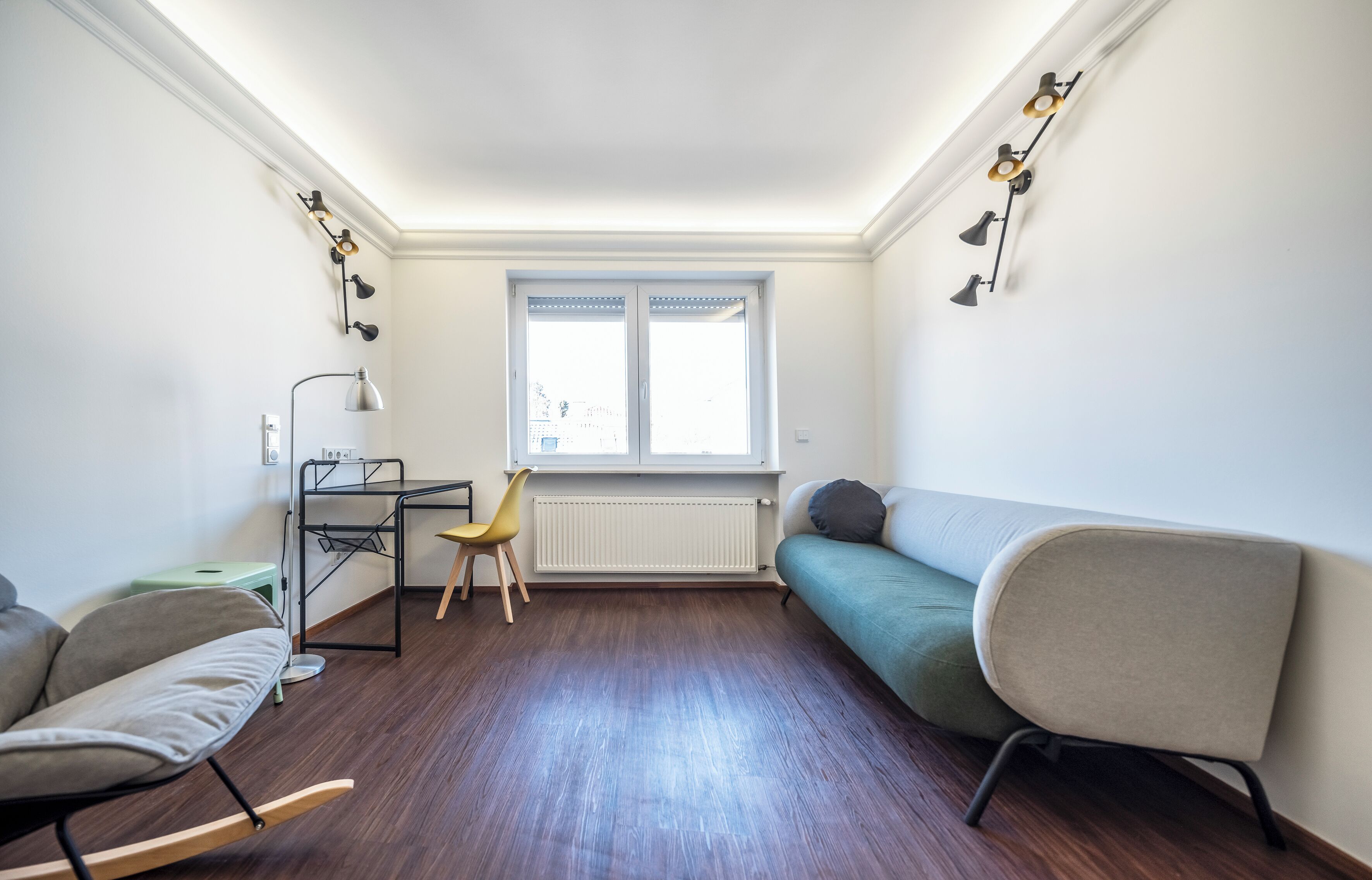 Et værelse med trægulv, en grøn sofa, en gul stol, et skrivebord med en lampe og en radiator.
