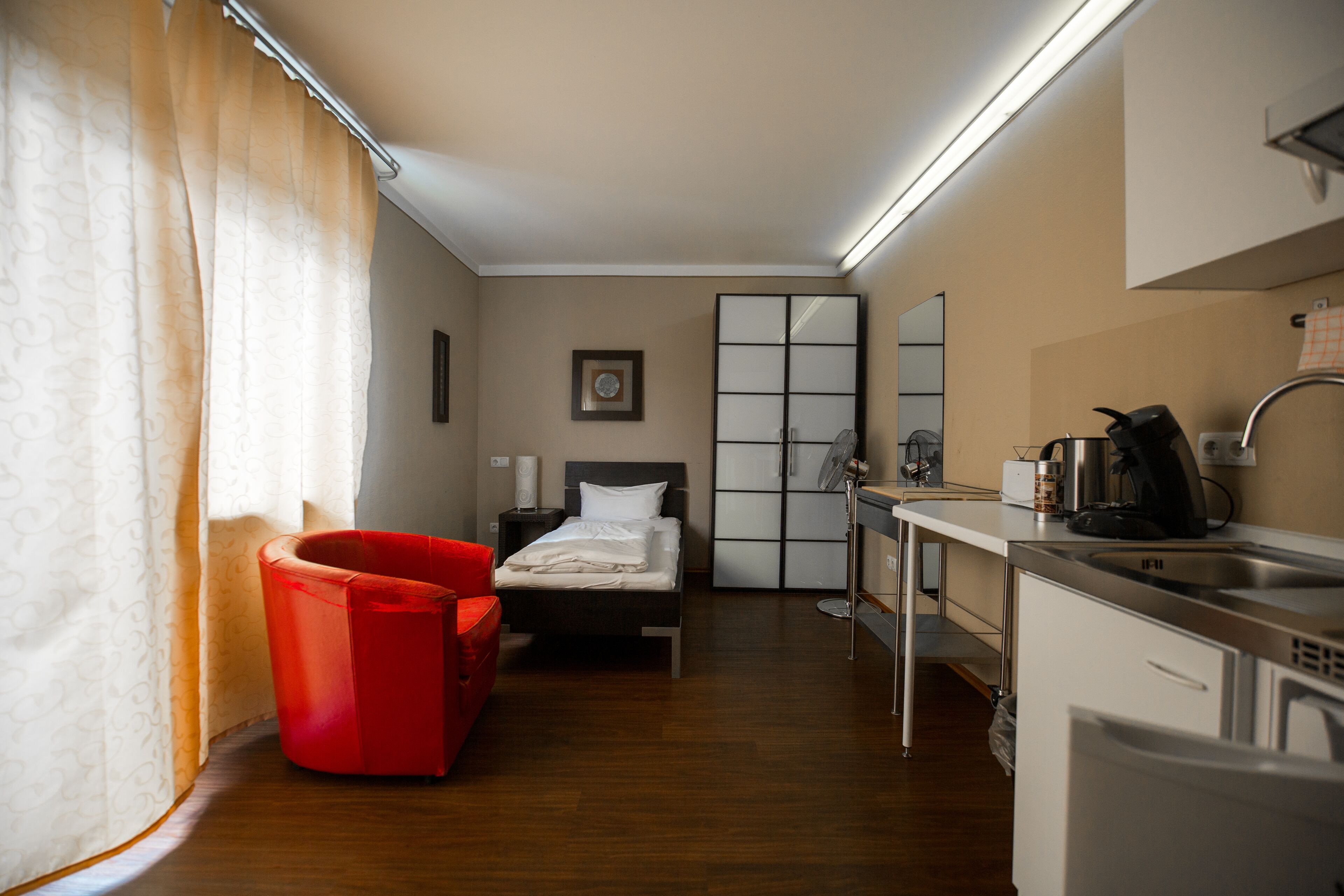 Ein kleines Hotelzimmer mit Bett, einem roten Sessel, einem kleinen Schreibtisch mit Wasserkocher und einer Küchenzeile.