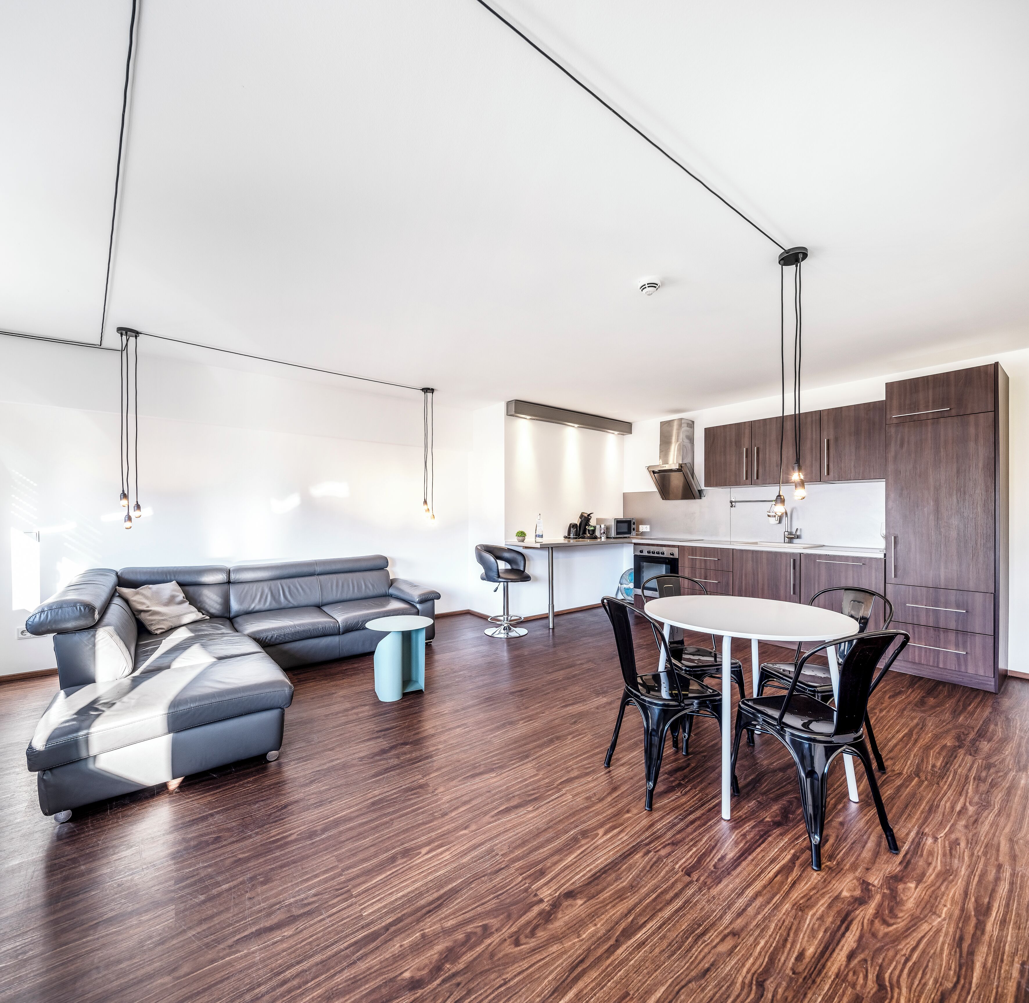 A modern living room with a sectional sofa, a dining table with chairs, and a kitchen area with wooden cabinets and a stainless steel range hood.