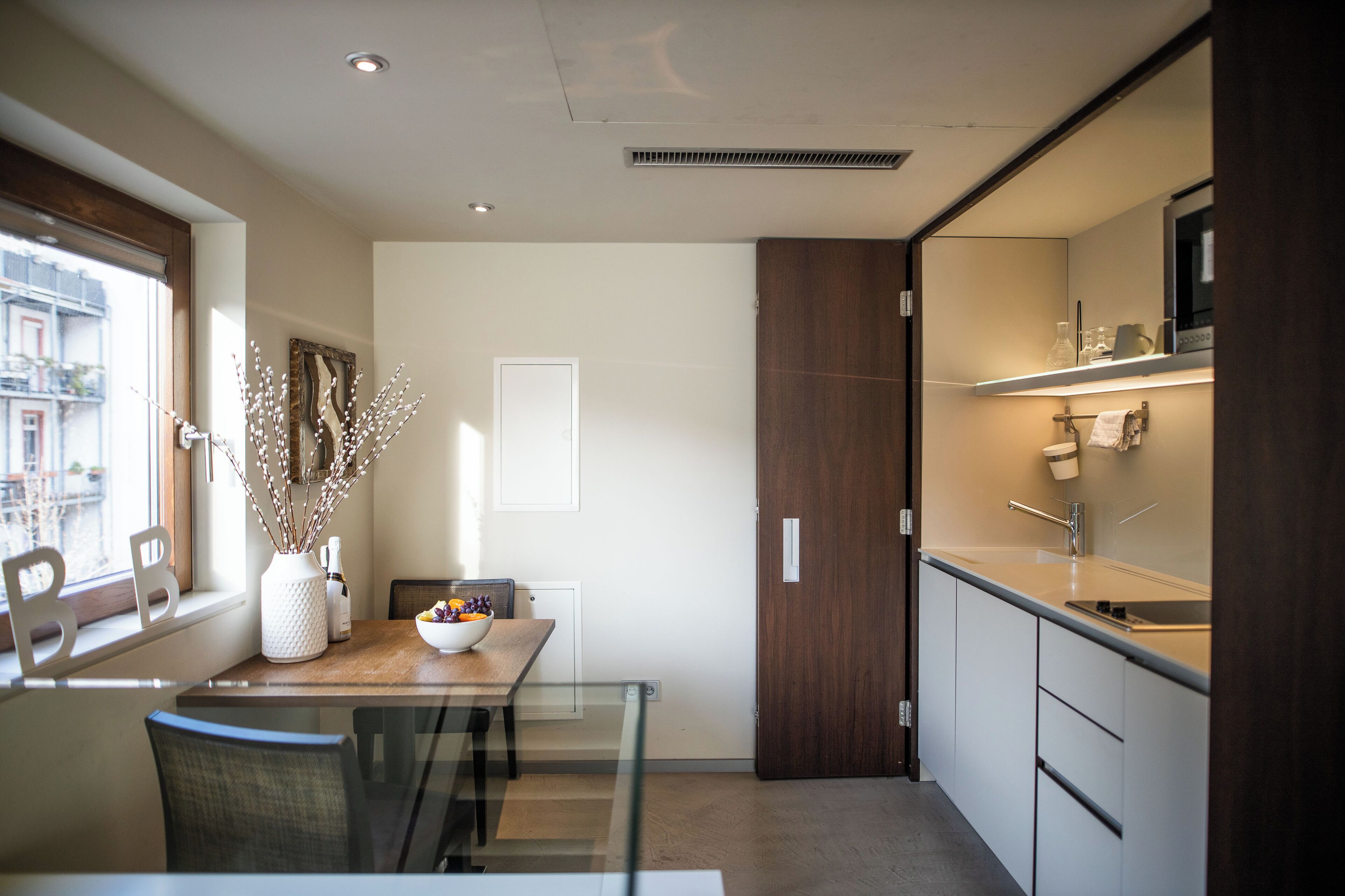 Una cocina moderna con zona de comedor, que incluye una mesa de madera, sillas, un florero con ramas y vistas a la ciudad a través de una ventana.