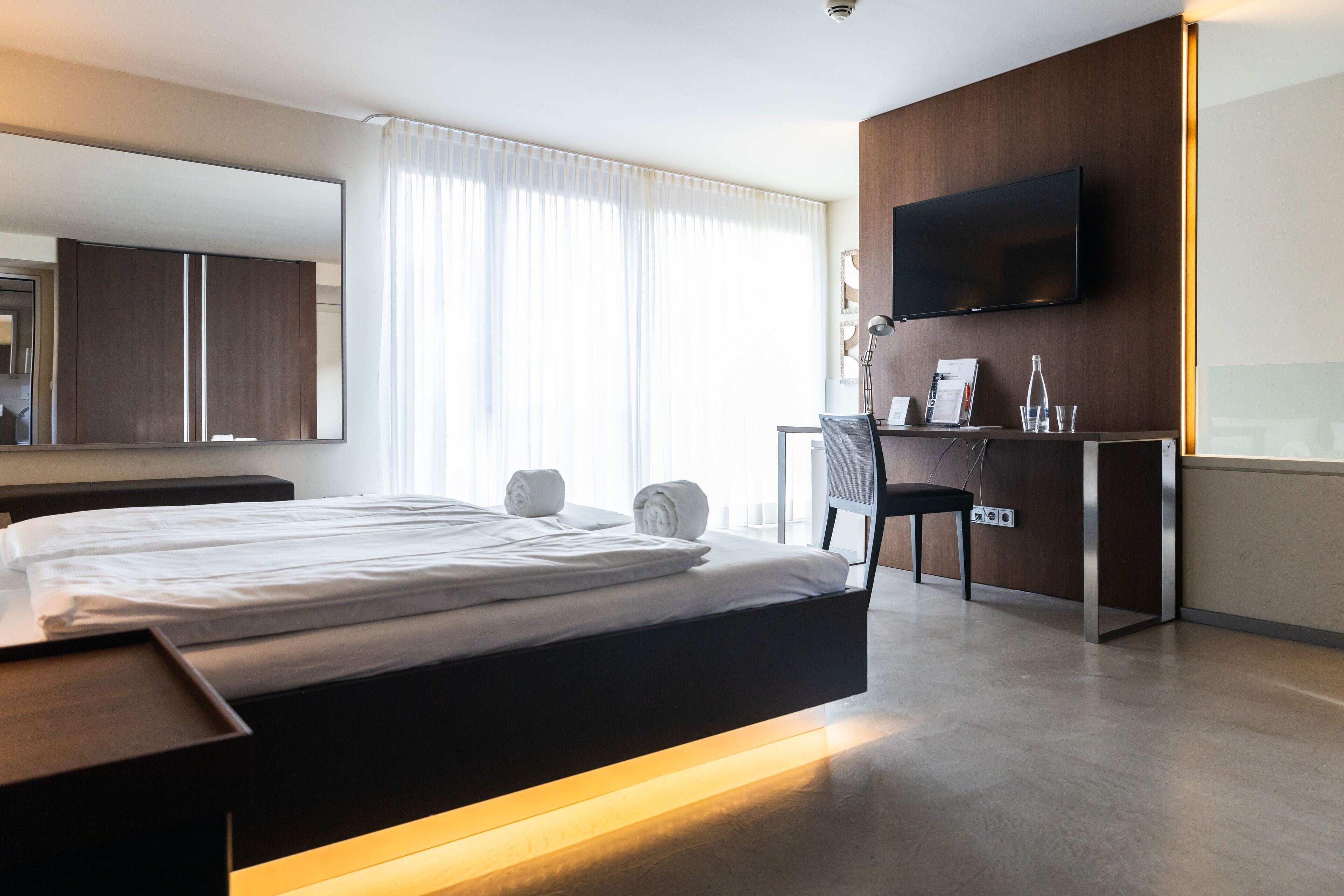 Ein modernes Hotelzimmer mit einem großen Bett, einem Schreibtisch mit Stuhl, einem Flachbildfernseher und einem großen Spiegel.