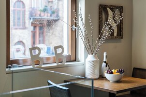 Un comedor con una mesa de madera, un cuenco de fruta, un florero con ramas y una botella de vino.
